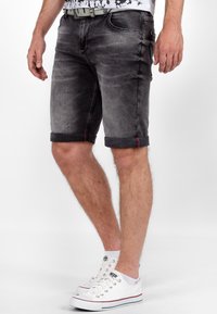 Heren grijze denimshorts met opgerolde manchetten, met subtiele slijtage, vijf zakken en rode stiksels, gecombineerd met witte sneakers.