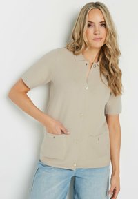Cardigan beige à manches courtes avec un col, boutonnière à l'avant et deux poches avant, fabriqué en matériau texturé doux.
