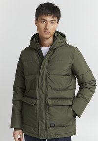 Puffjakke i mørkegrønn, med hette, glidelås, sidelommer og kviltet tekstur. Stoffet ser glatt ut og er vannavstøtende.