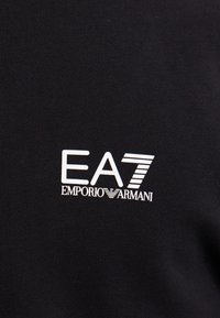 Fekete anyag textúrált felülettel, fehér "EA7 EMPORIO ARMANI" logóval, modern betűtípussal, egyszínű háttérrel.