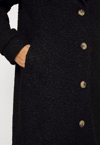 Cappotto nero testurizzato con grandi bottoni e una tasca laterale, realizzato in un materiale morbido e accogliente con un design a vestibilità ampia.