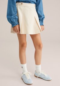 Helle beige Faltenrock aus Stoff, mit hoher Taille und Knöpfen. Kombiniert mit blauen Turnschuhen und weißen gerippten Socken.