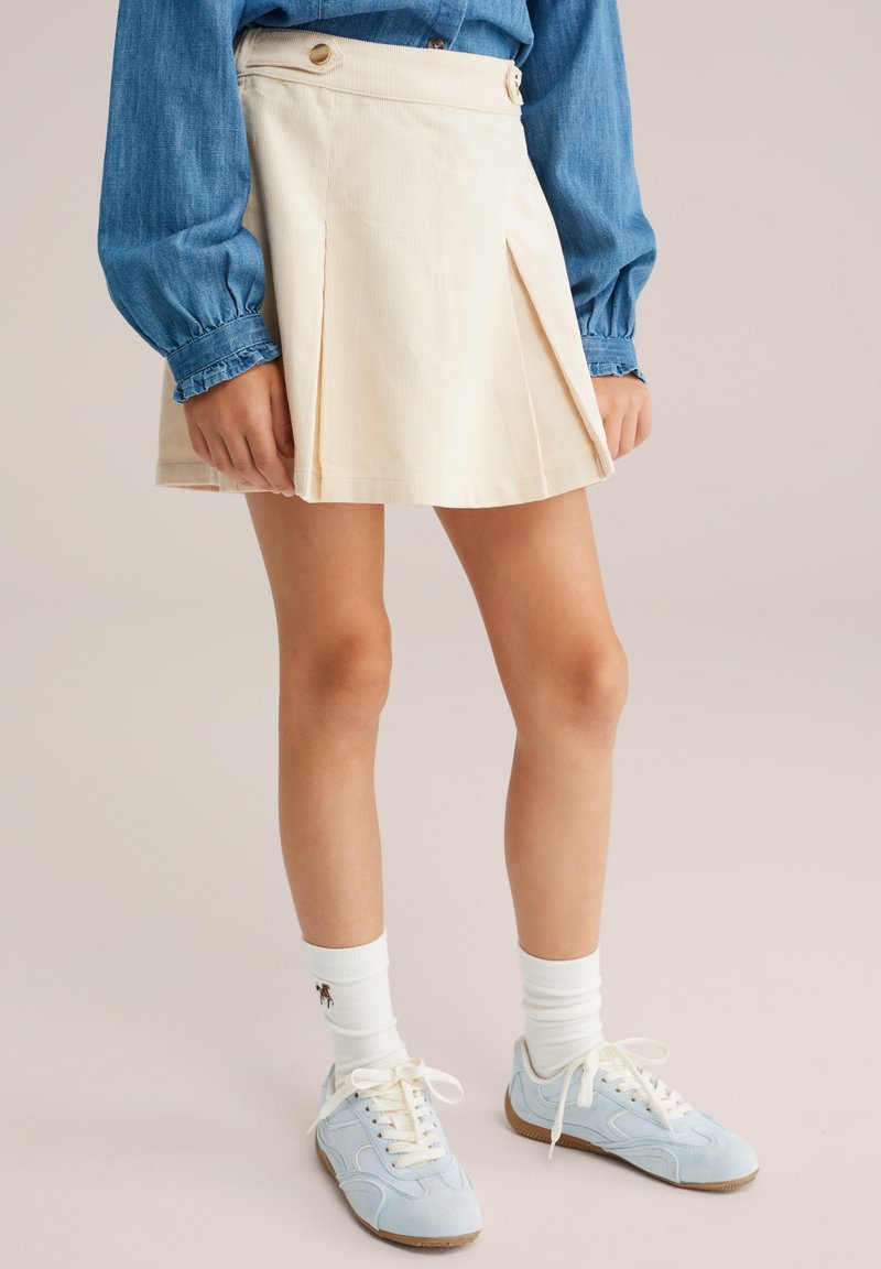Helle beige Faltenrock aus Stoff, mit hoher Taille und Knöpfen. Kombiniert mit blauen Turnschuhen und weißen gerippten Socken.