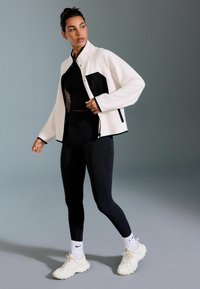Weiße und schwarze Zip-Jacke, schwarzes Cropped-Top, dunkle Leggings und weiße Sneakers mit strukturierten Sohlen. Glattes Material und lockere Passform.