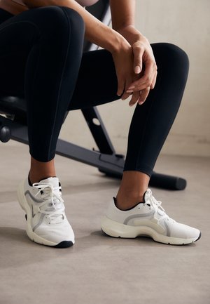 Person in black leggings sitting on exercise bench wearing white athletic sneakers on a gray floor.