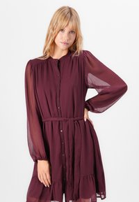 Burgundy, button-up dress with long, sheer sleeves, gathered bodice, and a ruffled hem. Includes a fabric belt for adjustable fit.