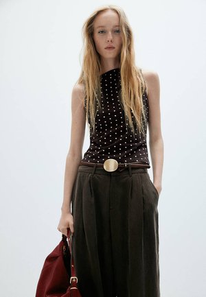Jeune femme aux longs cheveux blonds porte un top noir sans manches à pois, un pantalon foncé taille haute avec une grande boucle de ceinture ronde, tenant un sac rouge.