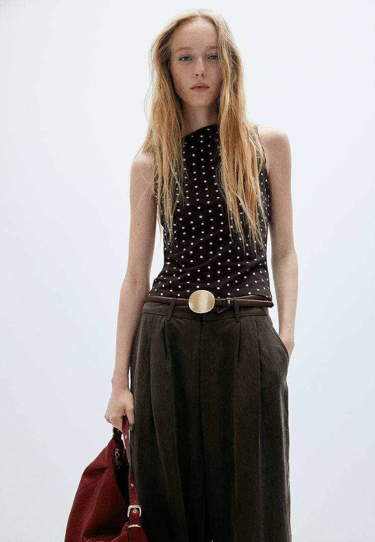 Jeune femme aux longs cheveux blonds porte un top noir sans manches à pois, un pantalon foncé taille haute avec une grande boucle de ceinture ronde, tenant un sac rouge.