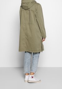 Oliwkowy płaszcz z kapturem o prostym kroju, z zamkiem błyskawicznym po bokach i widocznymi szwami. Nosi się go na jasnoniebieskie dżinsy z postrzępionymi nogawkami oraz białe sneakersy.