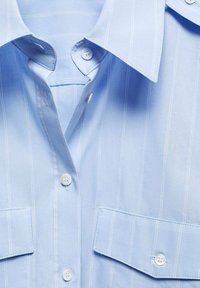 Chemise habillée bleu clair à rayures blanches verticales, boutonnée sur le devant, col pointu, avec deux poches poitrine à rabats et boutons.