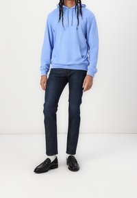 Personne portant un sweat à capuche bleu clair avec le logo GAP, un jean slim sombre, des chaussettes blanches et des mocassins en cuir noirs, se tenant devant un fond blanc uni.