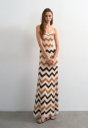 Femme debout portant une robe longue au crochet sans manches avec des motifs en zigzag marron, beige et blanc et des sandales noires, devant un mur uni.
