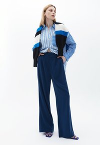 Model draagt een blauw-wit gestreept overhemd met knopen, gecombineerd met een wijdvallende marineblauwe broek en blauwe open teen sandalen, met een kleurbloktrui over de schouders gedrapeerd.