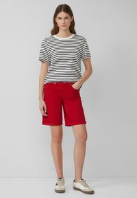 Bermuda en coton rouge avec des poignets retroussés, accompagnée d'un t-shirt à manches courtes noir et blanc à rayures. Modèle portant des baskets blanches et beiges.