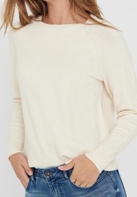 Heller cremefarbener Strickpullover mit Rundhalsausschnitt, Raglanärmel, gerippten Bündchen und Saum; kombiniert mit blauen Jeans. Weiche Textur, lässiger Schnitt.