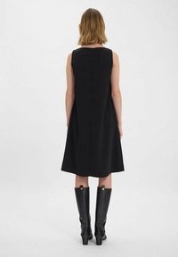Robe noire sans manches avec une silhouette en A, détail de fermeture éclair à l'arrière, portée avec des bottes en cuir noires montantes, texture lisse.