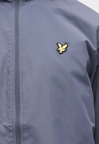 Light blue jacket made of smooth fabric, featuring a center zip and a small embroidered eagle logo in yellow and black.