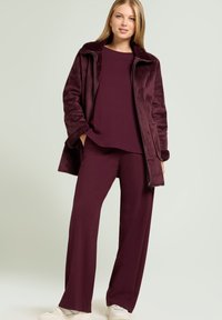 Chaqueta de forro polar burdeos con ribete de piel sintética, combinada con una parte superior y pantalones holgados burdeos, usados con zapatillas blancas. Texturas suaves en todo.