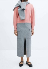 Personne portant un pull rose avec un pull gris drapé sur les épaules, une jupe midi grise avec une fente frontale et des poches à rabat, et des chaussures Mary Jane noires.