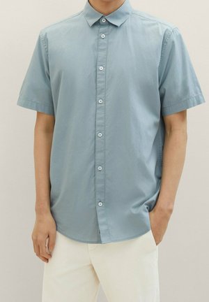 Man die een lichtblauw overhemd met korte mouwen en witte broek draagt, staand tegen een effen beige achtergrond.