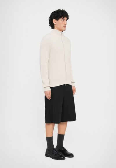 Κρεμ πουλόβερ με φερμουάρ και ριμπ ύφανση, συνδυασμένο με μαύρο σορτς μέχρι το γόνατο, γκρι κάλτσες και μαύρα παπούτσια, στέκεται μπροστά σε απλό φόντο.