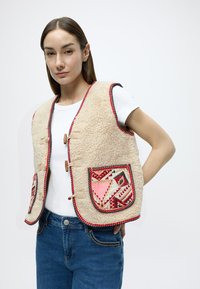 Μπεζ γιλέκο από συνθετική γούνα με κόκκινη επένδυση, με δύο τσέπες με σχέδιο και ξύλινα κουμπώματα. Φορώντας το πάνω από ένα λευκό πουκάμισο και μπλε τζιν.