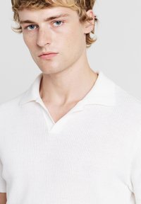 Giovane uomo con pelle chiara, capelli biondi e occhi azzurri, indossa una maglietta polo bianca in maglia, rivolto in avanti con un'espressione neutra.