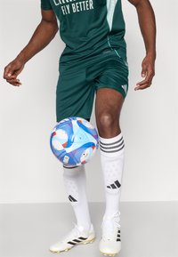 Vihreä urheilupaita, jossa valkoisella teksti "FLY BETTER", siihen sopivat shortsit, valkoiset jalkapallosukat mustin raidoin ja mustat nappikset. Värikäs jalkapallo on tasapainossa polven päällä.