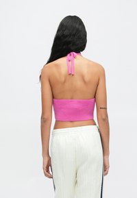 Top cropped halter em malha fúcsia com fecho amarrado no pescoço, combinado com calças texturizadas em cor creme com riscas laterais.