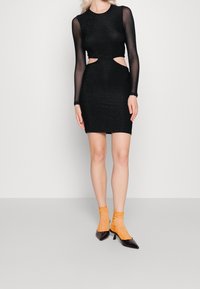 Černé glitrové šaty s dlouhými průhlednými rukávy, výřezy po stranách, přiléhavého střihu. V kombinaci s oranžovými texturovanými kotníkovými ponožkami a černými špičatými lodičkami.