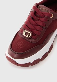Gros plan sur une sneaker bordeaux avec une semelle épaisse, des lacets rouges, un charme logo "G" en or et le logo Guess sur la languette.