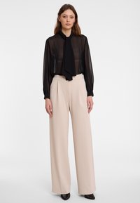 Zwarte transparante blouse met een halsstrik, gecombineerd met hoog-waist beige wijde broek. Glad materiaal, een ontspannen pasvorm en lange mouwen.