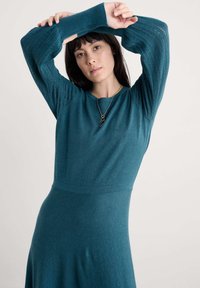 Mėlyna suknelė su ilgomis, tekstūruotomis rankovėmis ir prigludusia liemene, turinti apvalų iškirpimą ir subtilų išplatėjimą apatinėje dalyje.