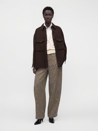 Femme portant une veste oversize marron foncé, une chemise crème, un pantalon large à carreaux et des talons pointus noirs, debout devant un fond blanc uni.