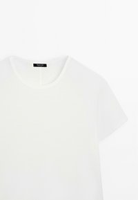 Hvid t-shirt med rund hals og korte ærmer, lavet af glat stof, med et sort mærke ved halsudskæringen. Ingen synlige mønstre eller detaljer.