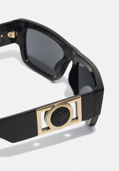Sorte solbriller med et dristig design og et gyldent rektangulært dekor, prydet med et løvehode-motiv, supplert med mørke linser.