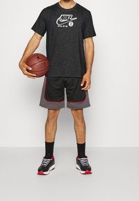 Hombre sosteniendo una pelota de baloncesto, vistiendo una camiseta negra de Nike, pantalones cortos de baloncesto negros y grises con ribetes rojos, calcetines negros y zapatillas negras con acentos rojos.