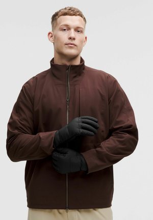 Junger Mann in brauner Jacke mit Reißverschluss, der schwarze isolierte Handschuhe trägt und einstellt, vor einfachem hellem Hintergrund.