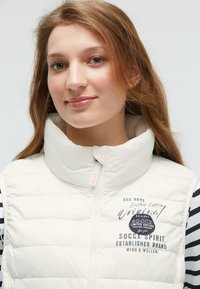 Weißer Steppwesten mit hohem Kragen, Reißverschluss und bedrucktem Logo sowie Text auf der Vorderseite. Gestreiftes Langarmhemd ist darunter sichtbar.