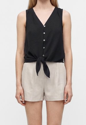 Femme portant un haut noir sans manches boutonné et noué à la taille, et un short beige clair taille haute, debout devant un fond uni.