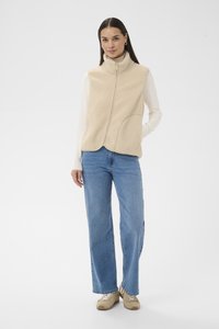 Femme portant un gilet en polaire beige sur un haut à manches longues blanc, un jean bleu et des baskets beiges, debout avec une main dans la poche.