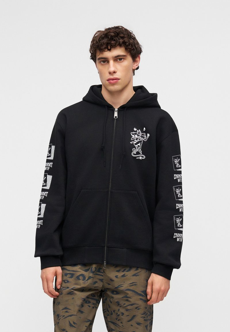 Junger Mann trägt einen schwarzen Hoodie mit Reißverschluss, weißen grafischen Designs und grünen Leoparden-Print-Hosen, steht vor einem einfarbigen hellen Hintergrund.