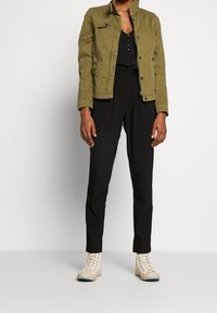 Olivově zelená bunda se strukturovaným designem, spárovaná s černým topem a zúženými černými kalhotami. Outfit doplňují béžové šněrovací boty.