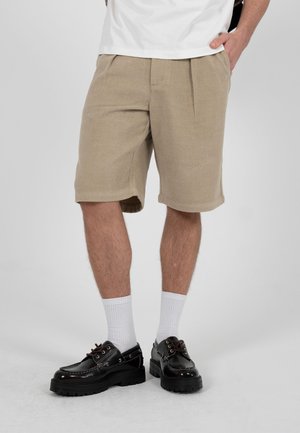 Man draagt beige kniebroek, witte sokken en zwarte leren veterschoenen tegen een effen grijze achtergrond.