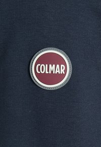 Námořnická modrá tkanina s kulatou gumovou nášivkou na vínovém pozadí a bílým nápisem "COLMAR", umístěná na texturovaném povrchu.
