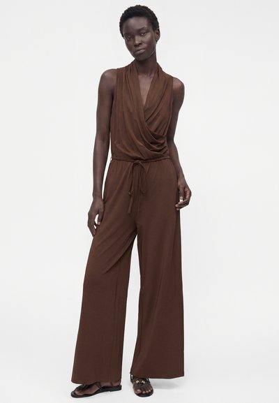 Femme portant une combinaison marron sans manches à jambes larges, avec un col drapé et une ceinture nouée à la taille, debout devant un fond clair uni.