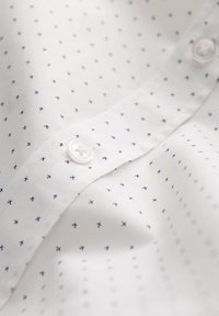 Camicia bianca con un piccolo motivo a croce blu, dotata di un colletto classico e una chiusura a bottoni. Il tessuto appare liscio e morbido.