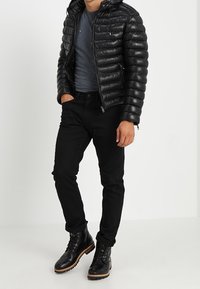 Zwarte gewatteerde jas met capuchon, grijs T-shirt eronder, zwarte spijkerbroek en zwarte laarzen met bruine zolen en veters. Casual winteroutfit.