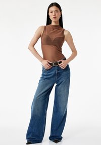 Bruine mouwloze top met een hoge hals en subtiele ronde naden, gecombineerd met losse blauwe denim jeans en zwarte puntige hakken.