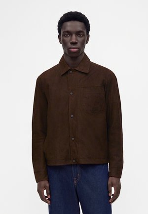 Junger schwarzer Mann trägt eine dunkelbraune Jacke mit Knöpfen und Brusttasche sowie blaue Jeans, steht vor einem schlichten hellen Hintergrund.